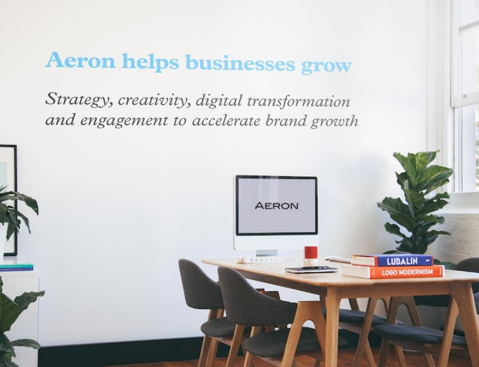 An Aeron meeting room wall carrying the line Aeron helps businesses grow, above a desk and design books.