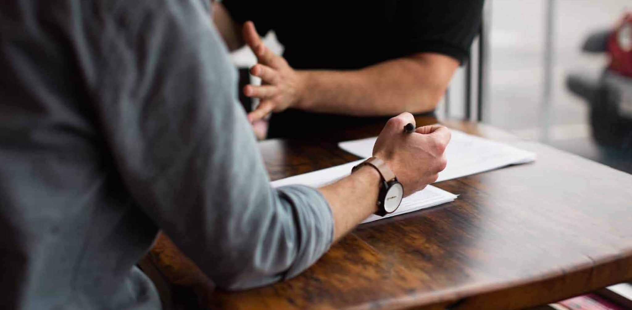 Two people in discussion across a desk, one taking notes, during a research interview.