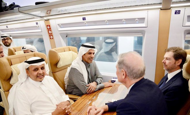 Passengers seated around a table in a Saudi Arabia Railways carriage, in conversation during a journey.