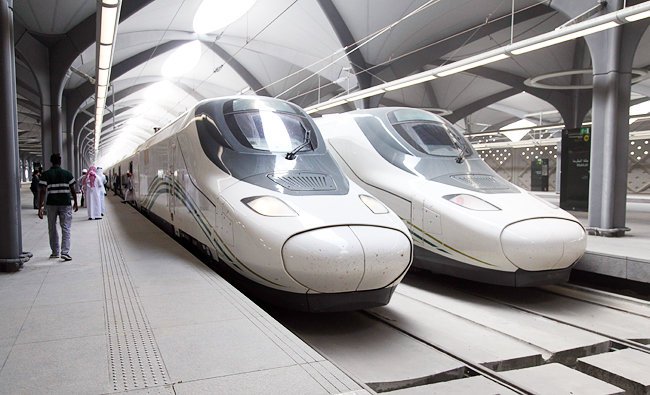 Two Haramain high speed trains stationary under the sculptural roof canopy of a Saudi railway station.