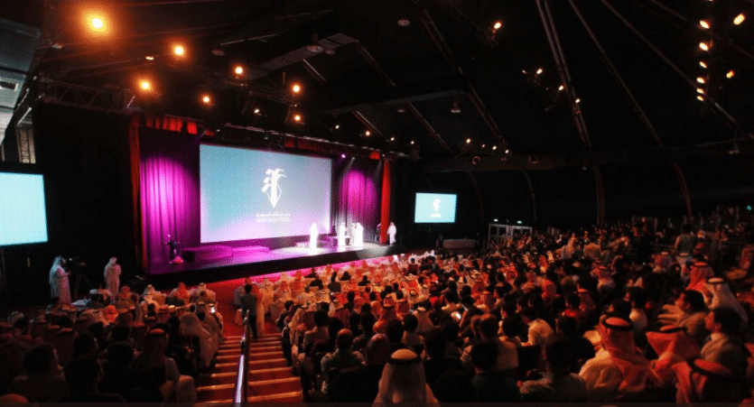 A packed auditorium in Saudi Arabia watching a screening, lit in pink and amber stage light.