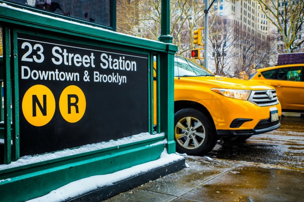 The 23 Street subway station sign in New York, with a yellow cab passing on the street behind.