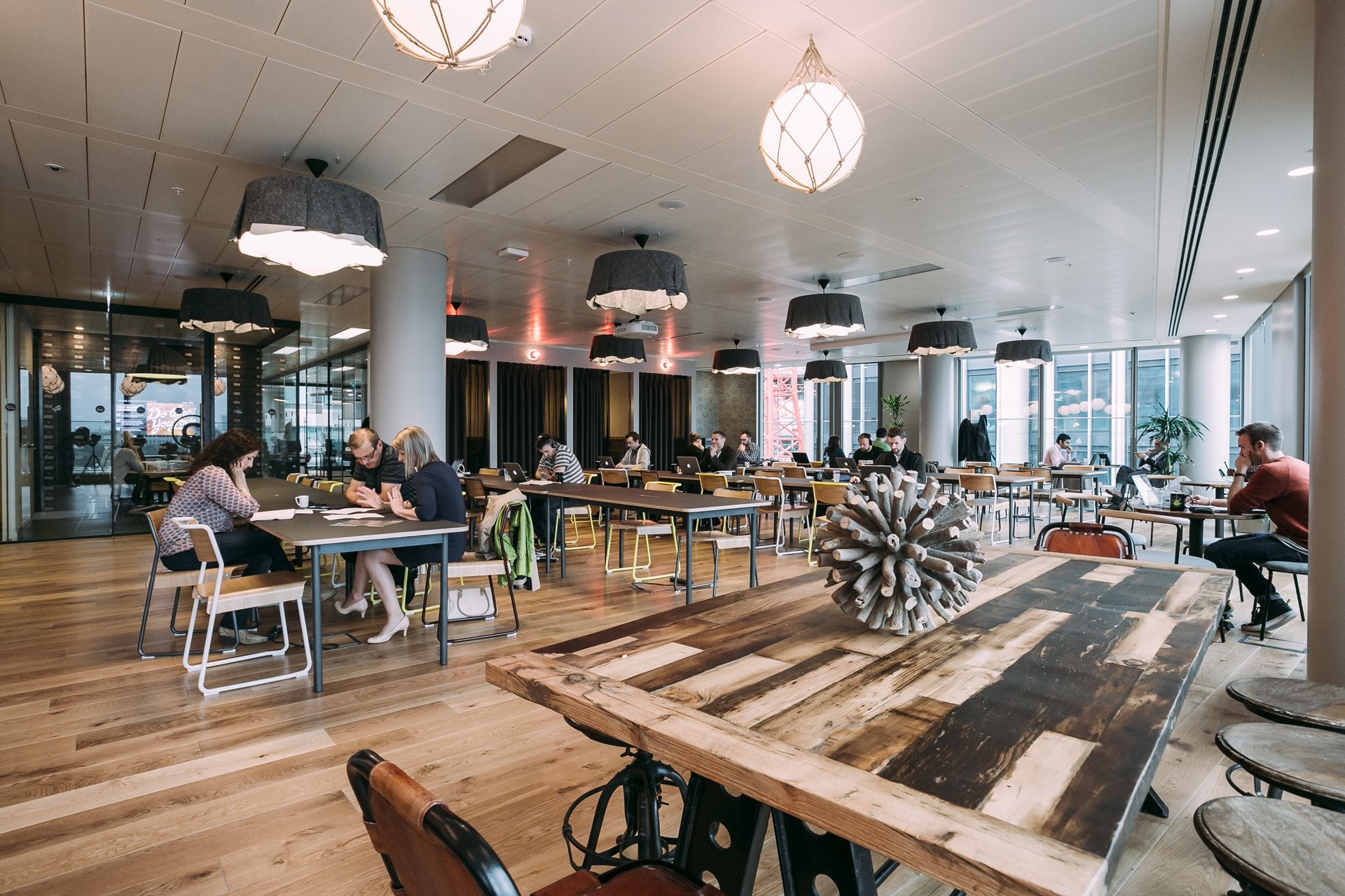 A WeWork co-working floor with long shared timber tables, pendant lighting and members working at laptops.