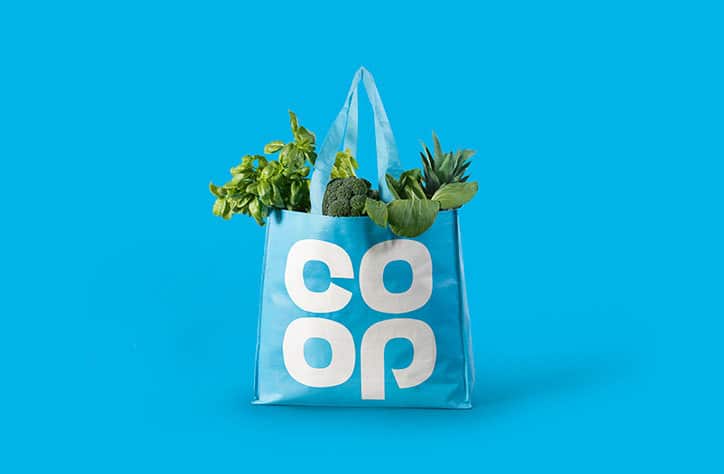 A Co-op reusable carrier bag in pale blue filled with fresh vegetables, carrying the redrawn Co-op cloverleaf.