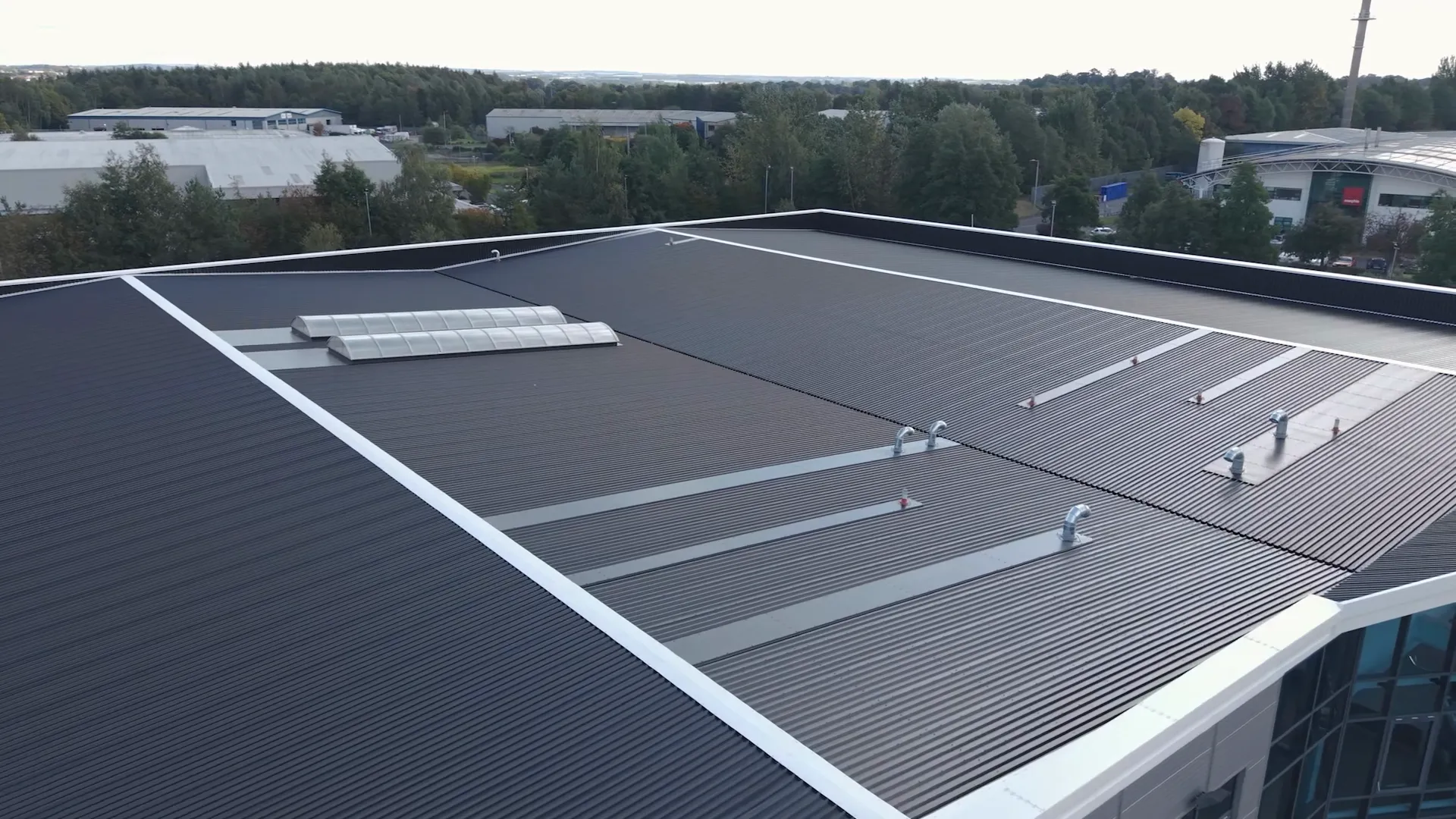 Aerial view of a large industrial building with a dark corrugated metal roof featuring vents and a small translucent skylight, surrounded by trees and other industrial buildings.