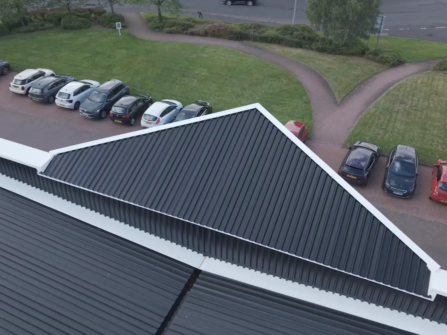 Aerial view of a building with a dark corrugated metal roof and white trim, with parked cars and green grass surrounding it.