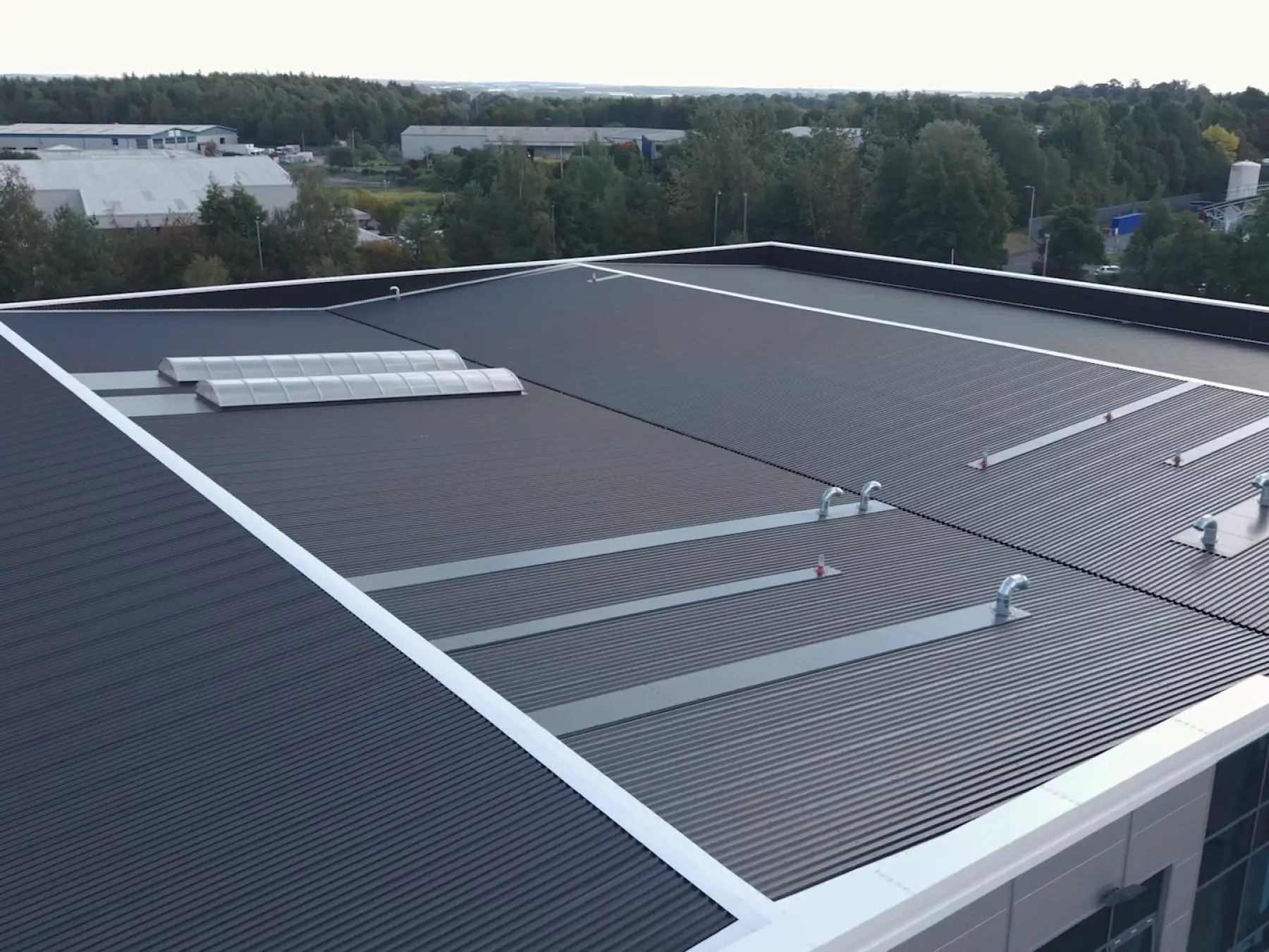 Aerial view of a large industrial building with a dark gray corrugated metal roof featuring multiple roof vents and two curved translucent rooflights, surrounded by trees and additional buildings in the background.