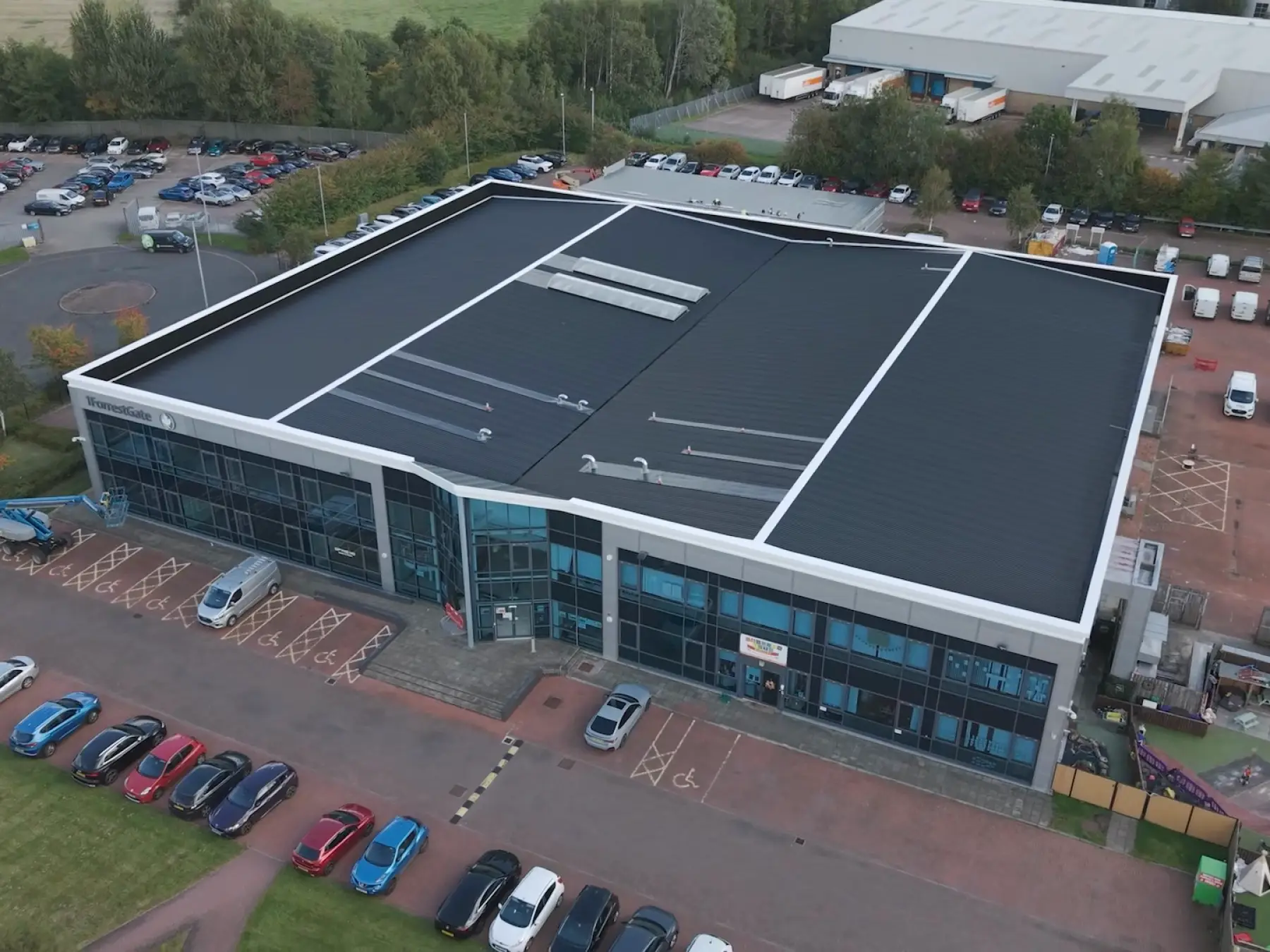 Aerial view of a modern office building with a black sectioned roof and large glass windows, surrounded by parked cars and greenery.