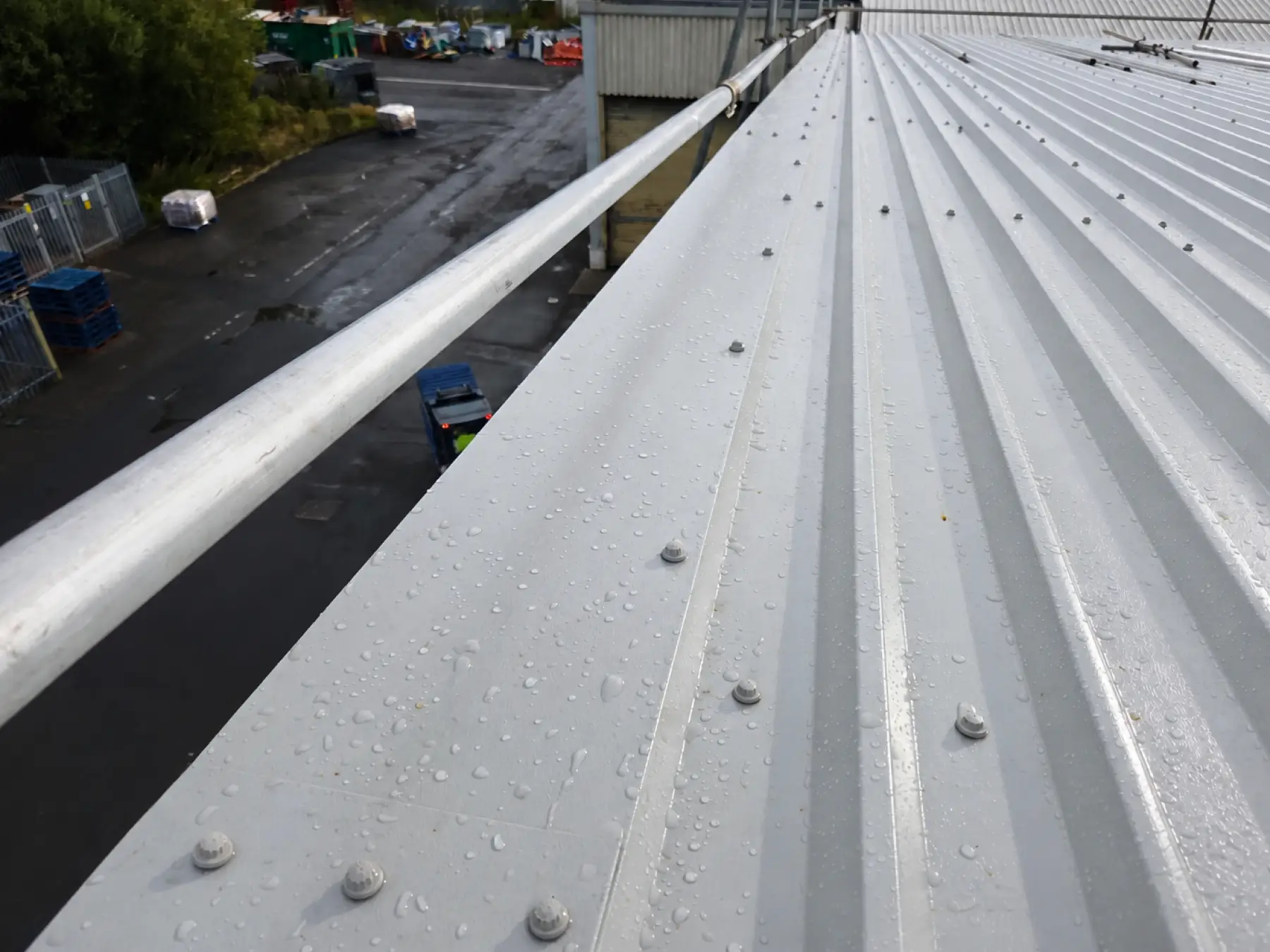 Wet gray metal roof with water droplets and bolts, viewed from a side railing, overlooking an industrial area with pallets and containers.