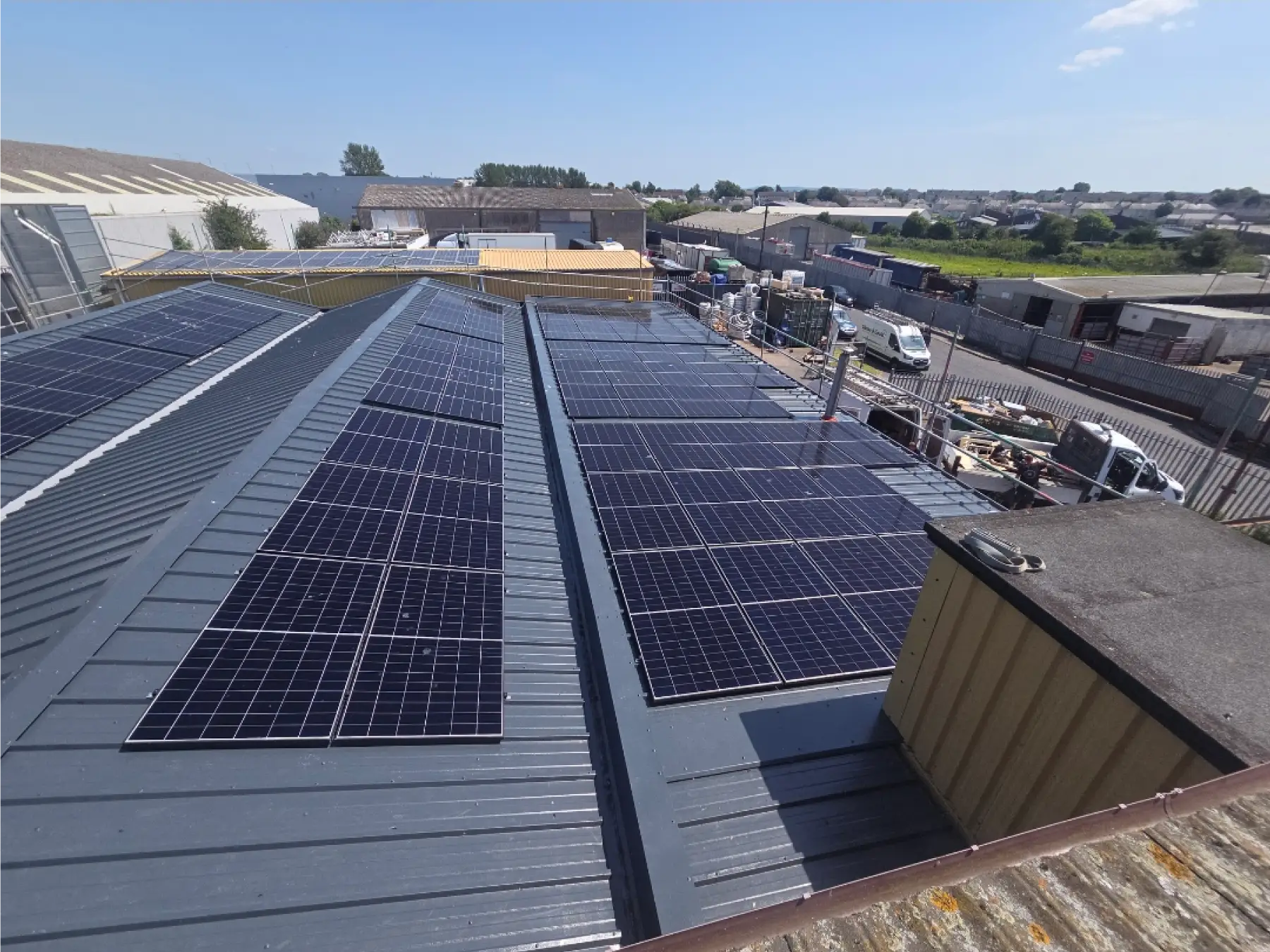 Solar panels installed on a large metal industrial rooftop under a clear blue sky, with industrial buildings and parked vehicles visible in the background.