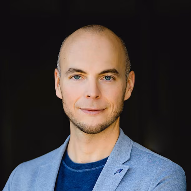 Man with short beard and blue eyes wearing a light gray blazer and navy shirt with black background.