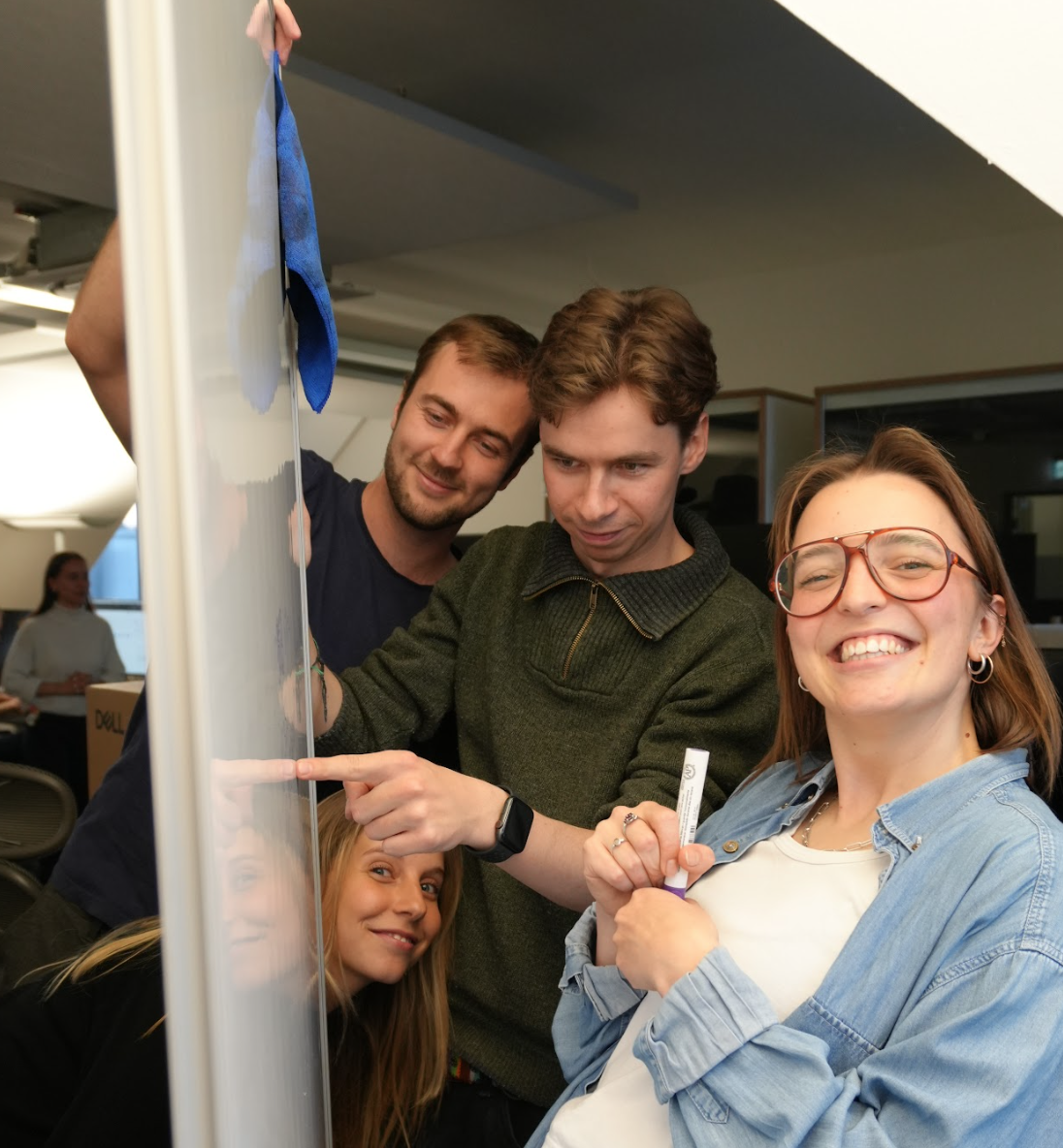Four smiling young adults collaborate at a whiteboard, one cleaning, two pointing, one holding a marker.