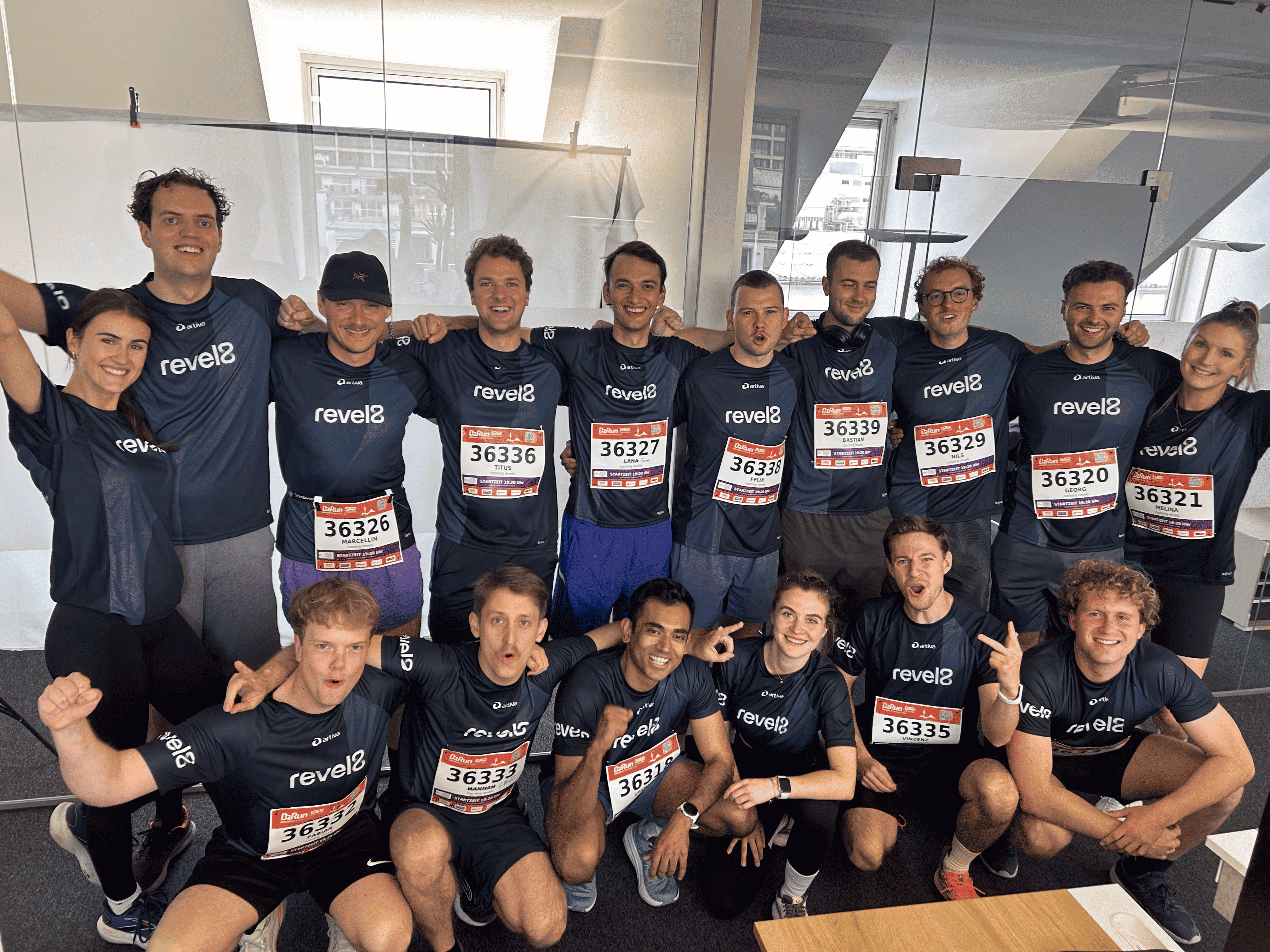 Group of 17 runners in matching dark revel8 shirts posing indoors with race bibs and smiling.