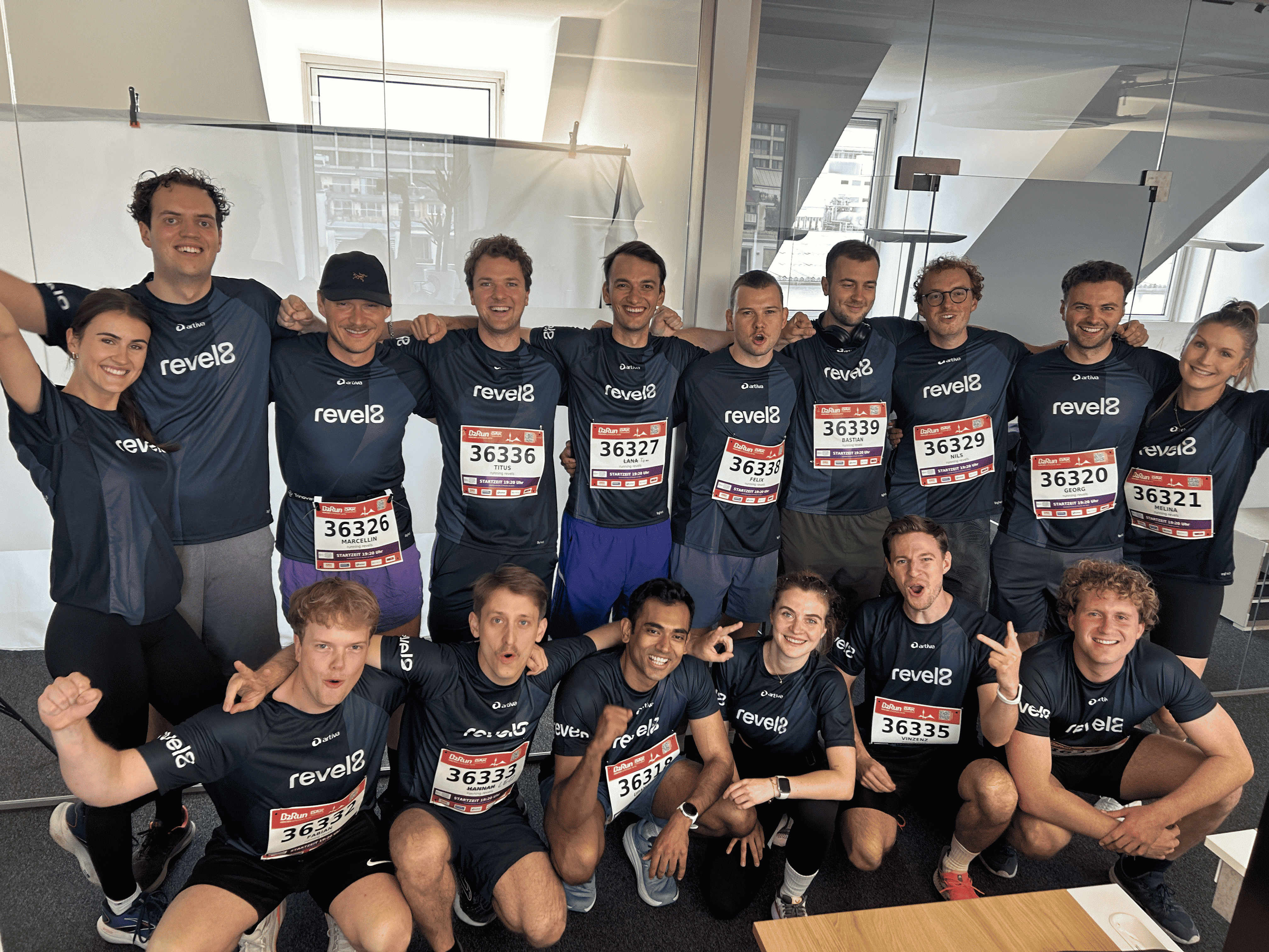 Group of 17 runners in matching dark revel8 shirts posing indoors with race bibs and smiling.