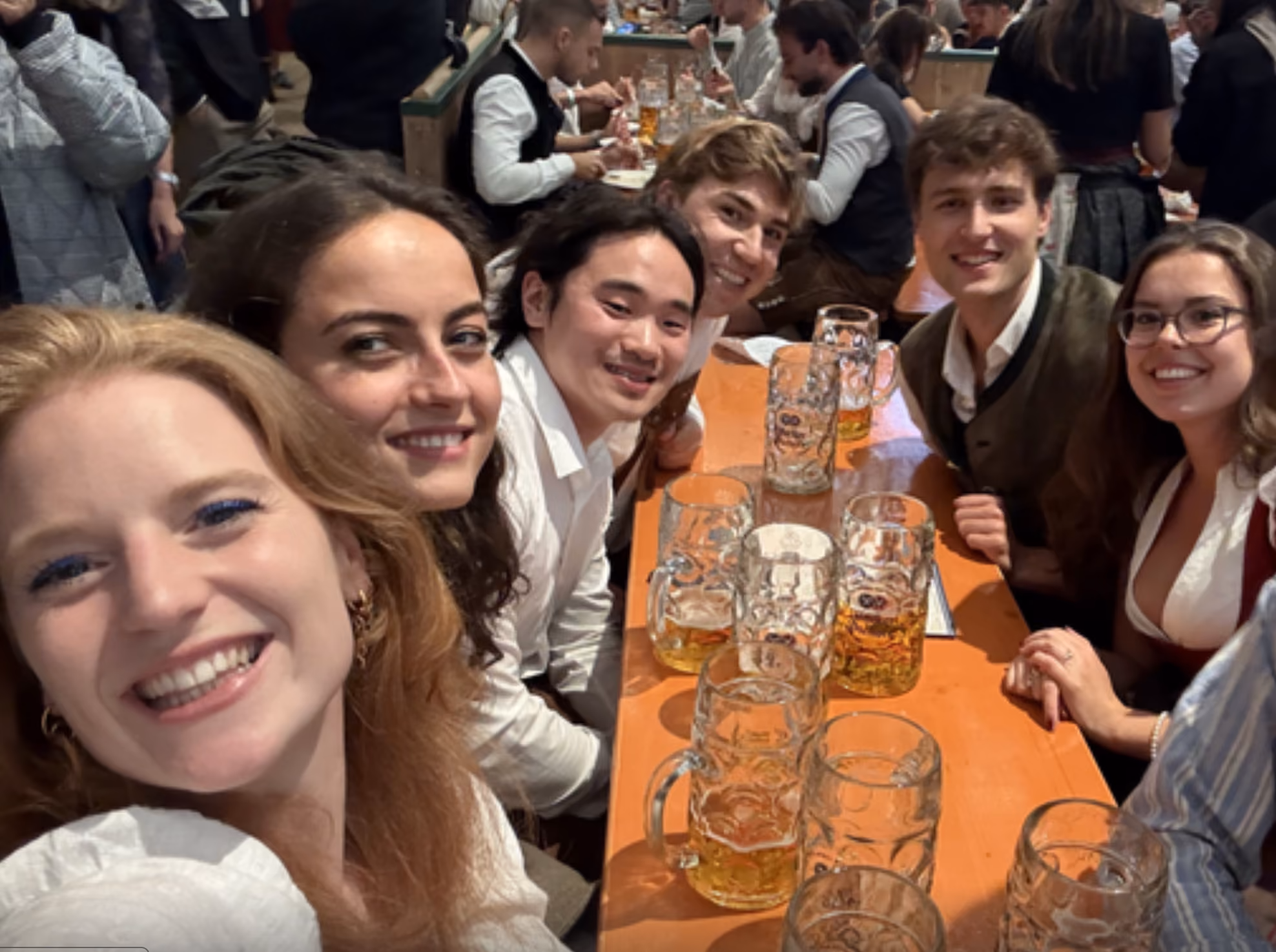 Group of six friends smiling at a table with large beer mugs at a crowded beer hall.