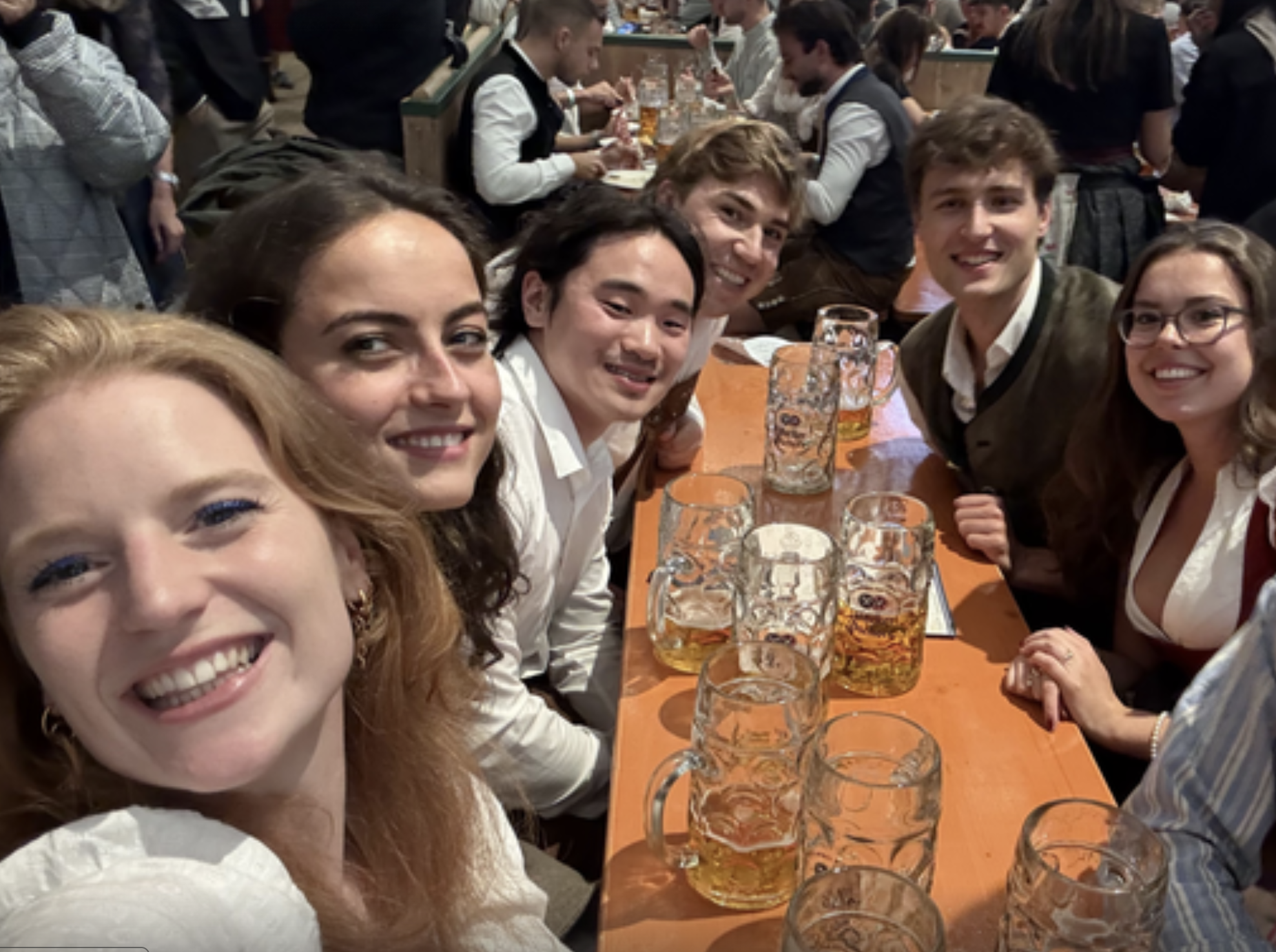 Group of six friends smiling at a table with large beer mugs at a crowded beer hall.