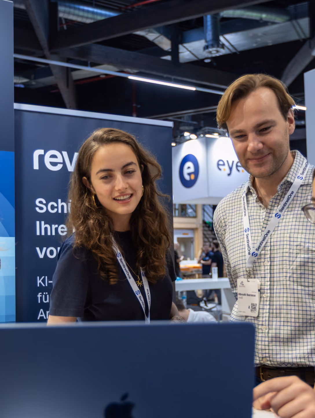 Two people with lanyards looking at a laptop at a tech event booth with branded banners.