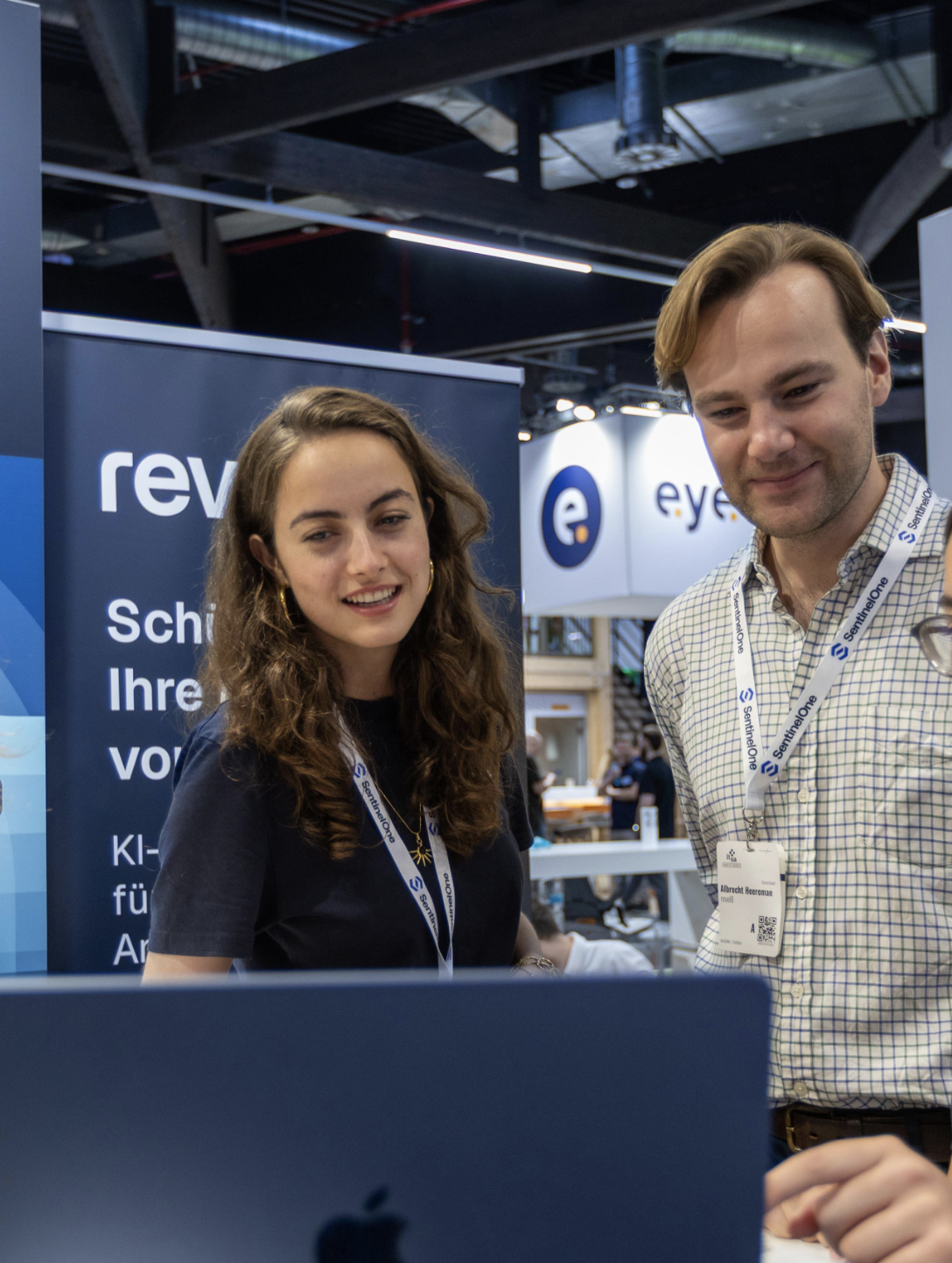 Two people with lanyards looking at a laptop at a tech event booth with branded banners.
