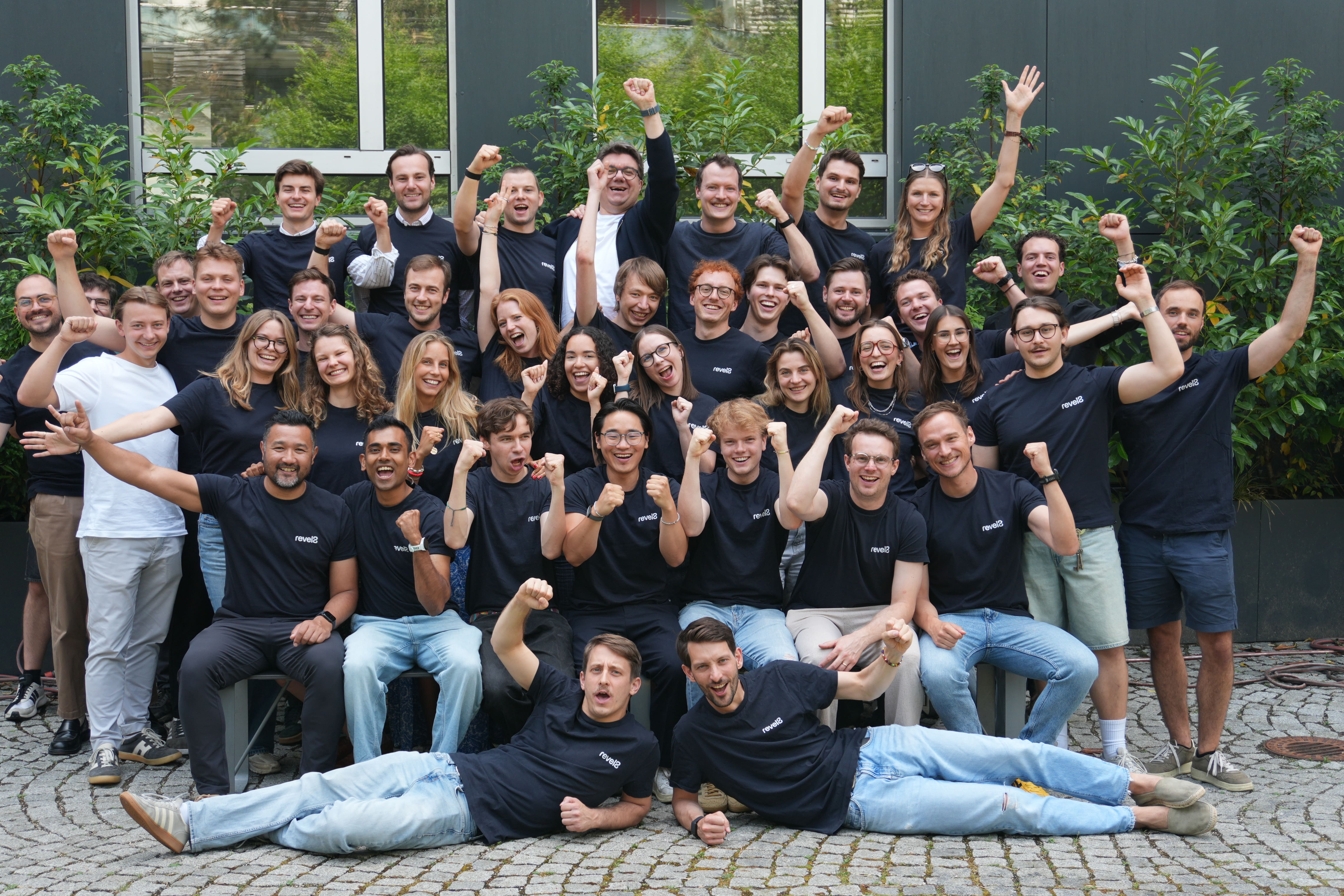 Large group of people in black and white shirts posing energetically with raised fists outdoors on stone pavement.