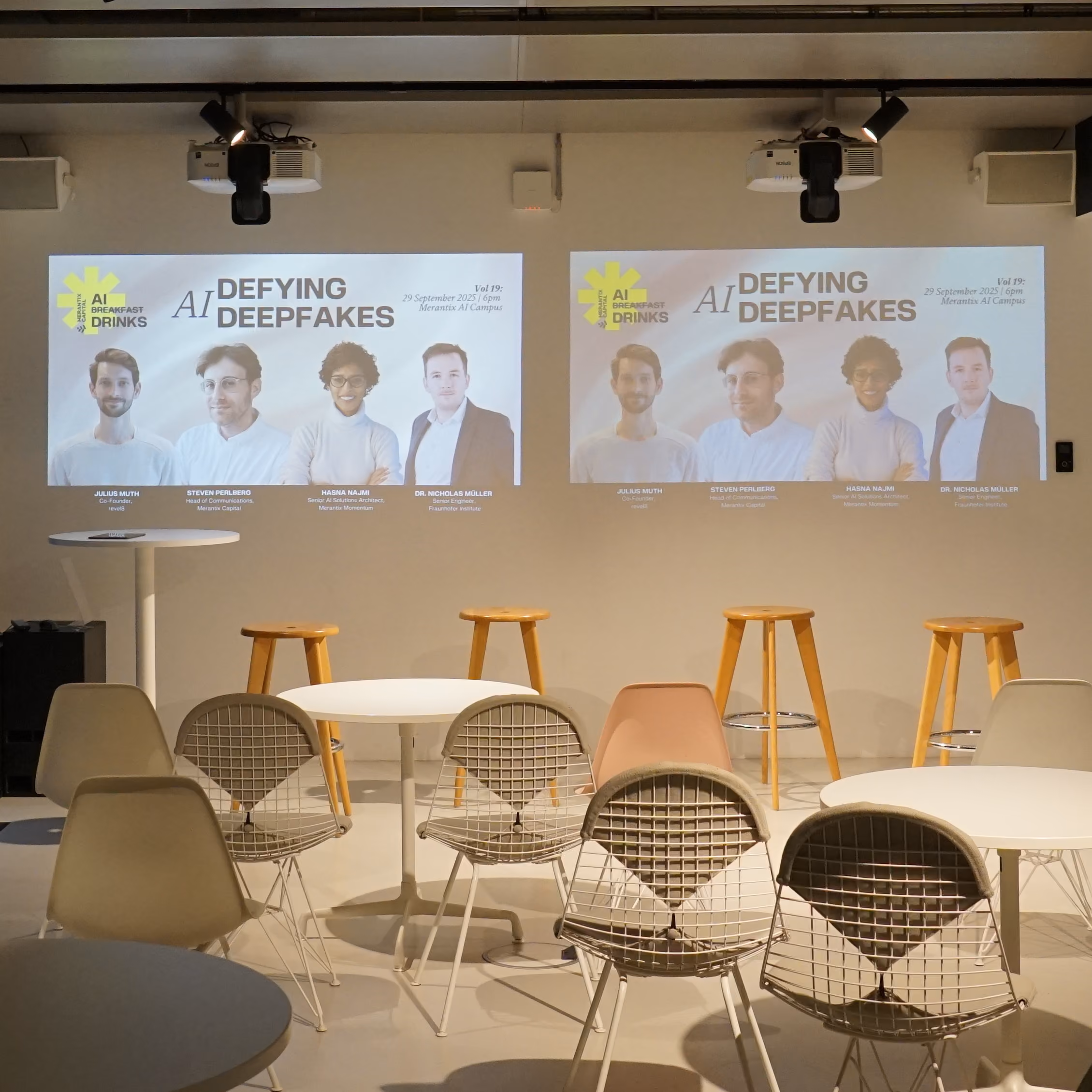 Empty event space with chairs and stools facing two screens showing AI Defying Deepfakes speaker info.