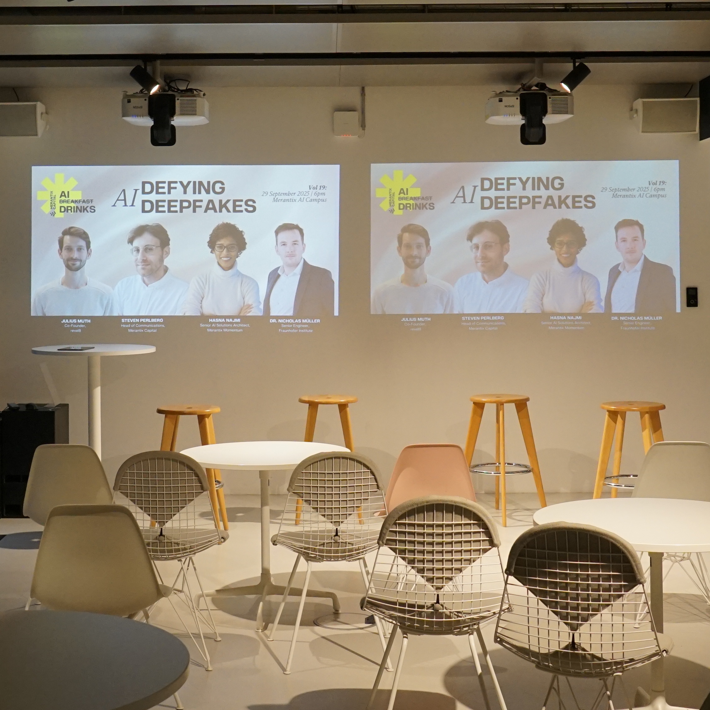 Empty event space with chairs and stools facing two screens showing AI Defying Deepfakes speaker info.