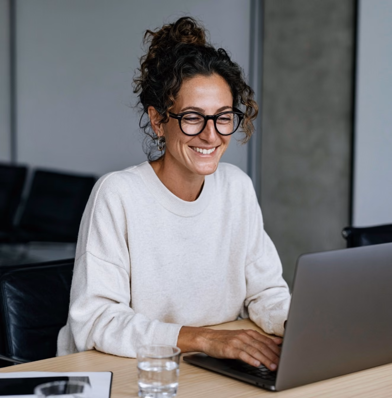 Smiling woman with glasses and curly hair working on a laptop at a wooden desk.
