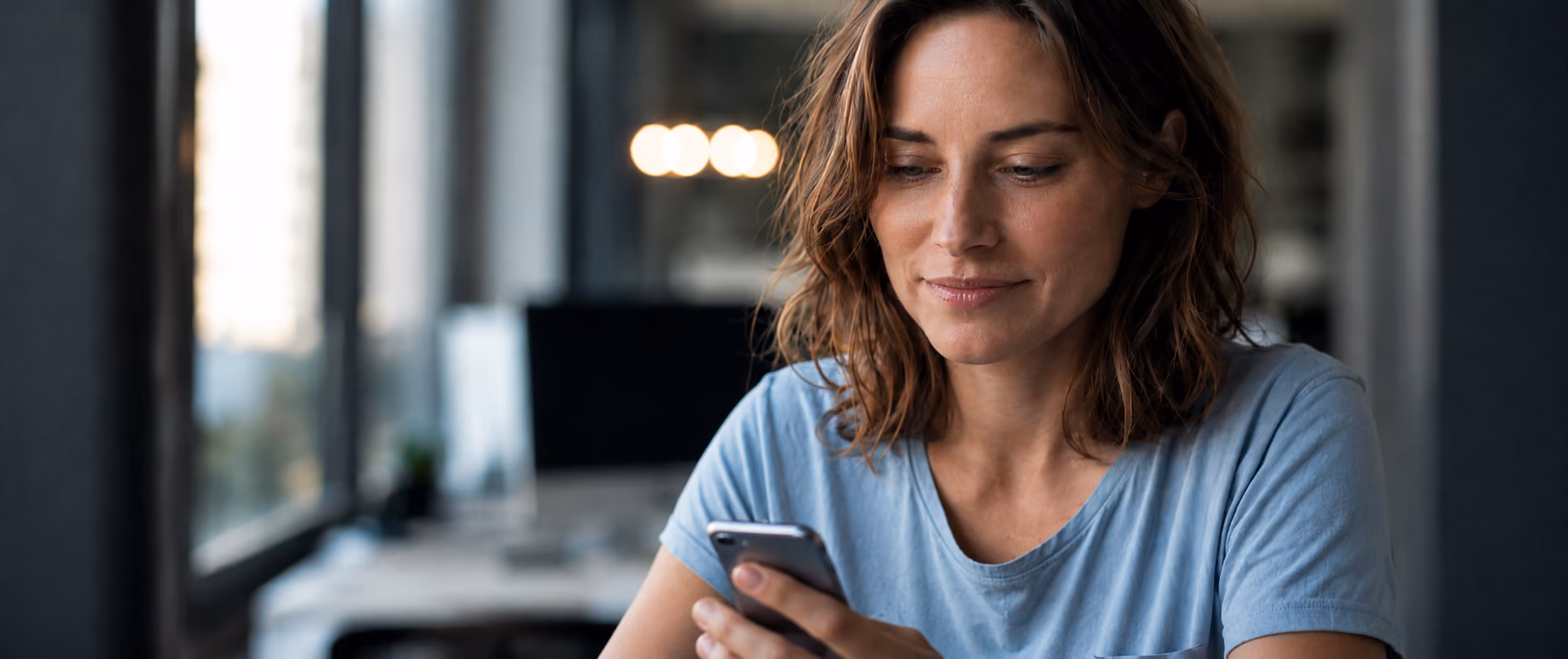 Woman in blue shirt looking at a smartphone with a slight smile in a blurred indoor setting.