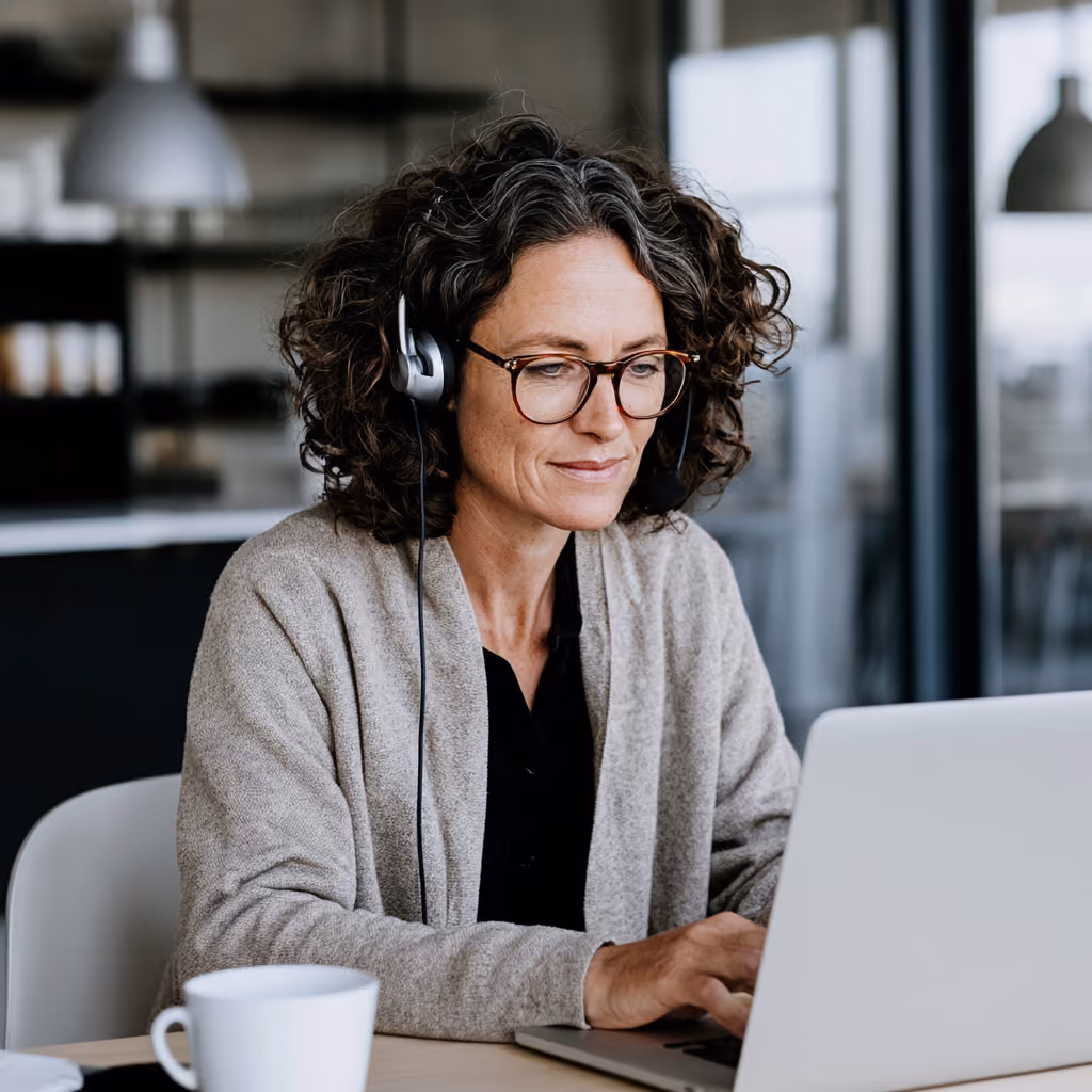 Woman with curly hair and glasses wearing headset, working on a laptop at a desk with a white mug.