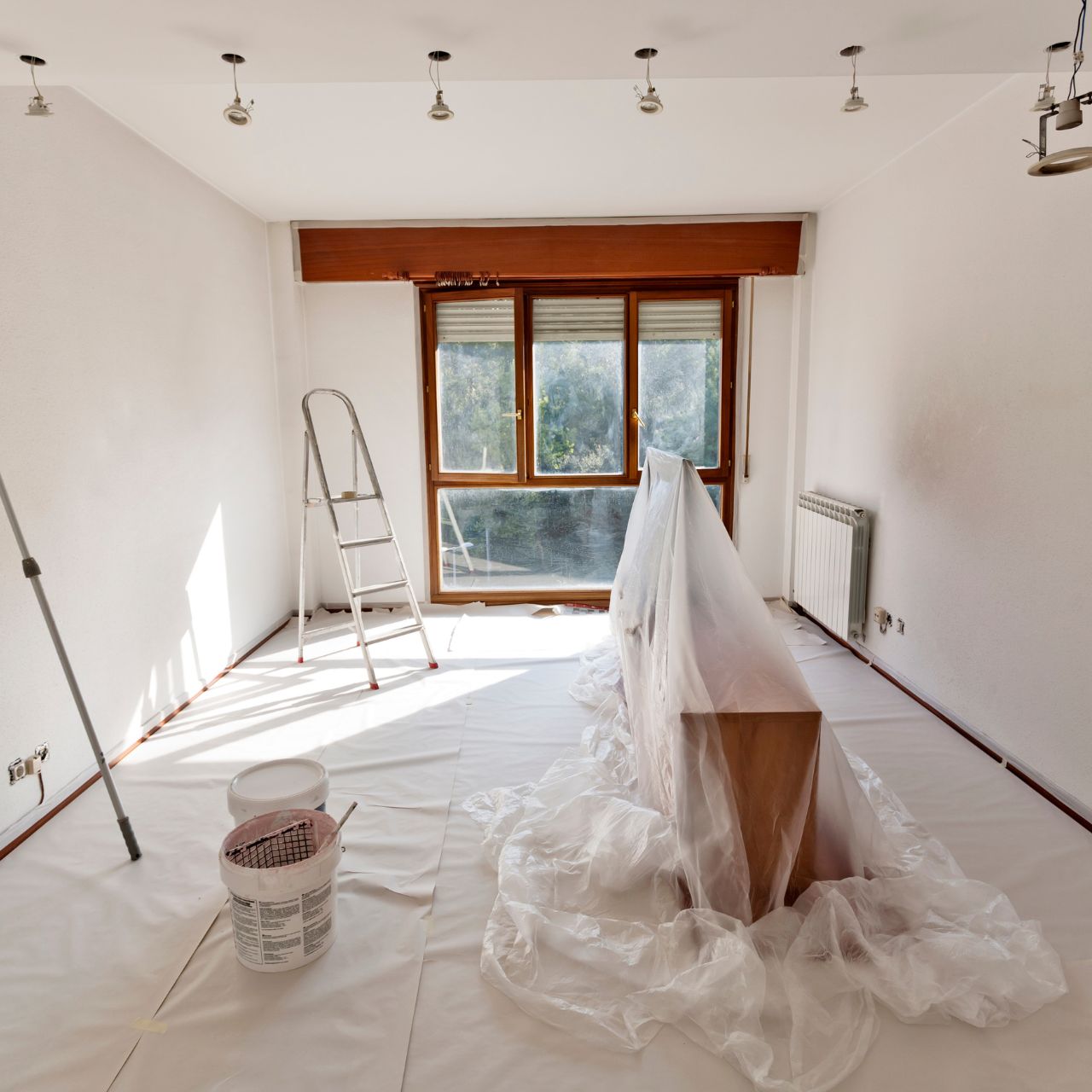Room mid-renovation with ladder, paint bucket, covered furniture, and large window letting in natural light.
