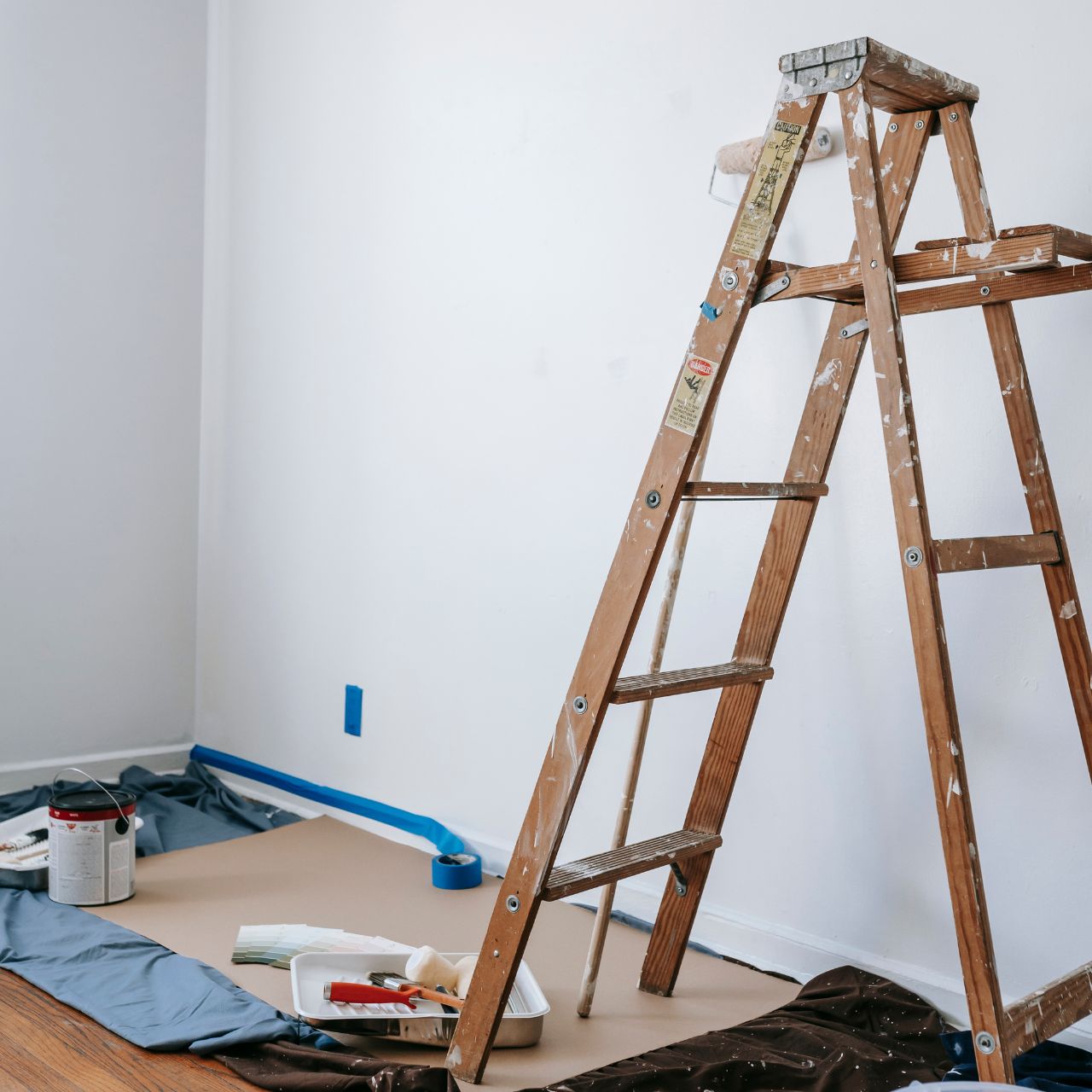 Wooden ladder with paint splatters by wall with painting supplies and drop cloth on floor.