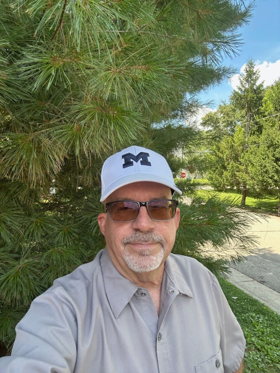 Middle-aged man with glasses, gray shirt, and white cap with M, standing outdoors near pine trees.