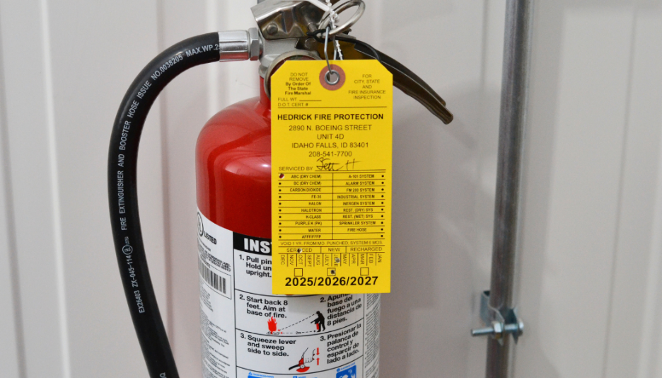 Red fire extinguisher with a yellow inspection tag attached, mounted on a wall with a metal pipe.