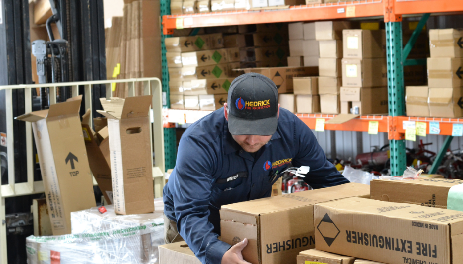 Warehouse worker in Hedrick Fire Protection uniform handling cardboard boxes labeled fire extinguisher.