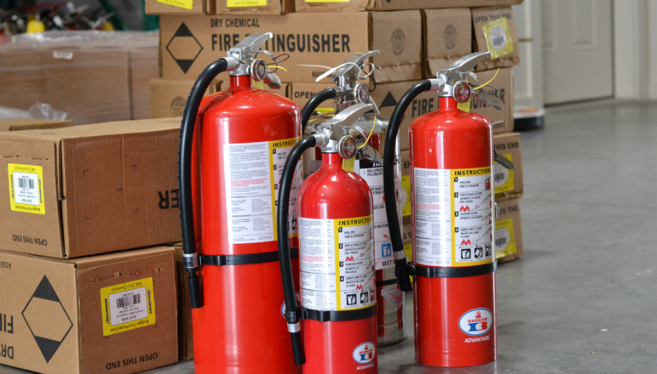 Four red fire extinguishers with black hoses standing on a warehouse floor near stacked boxes.