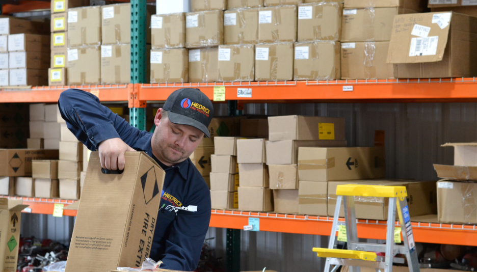Warehouse worker wearing a black cap handling a large dry chemical fire extinguisher box among stacked cardboard boxes.