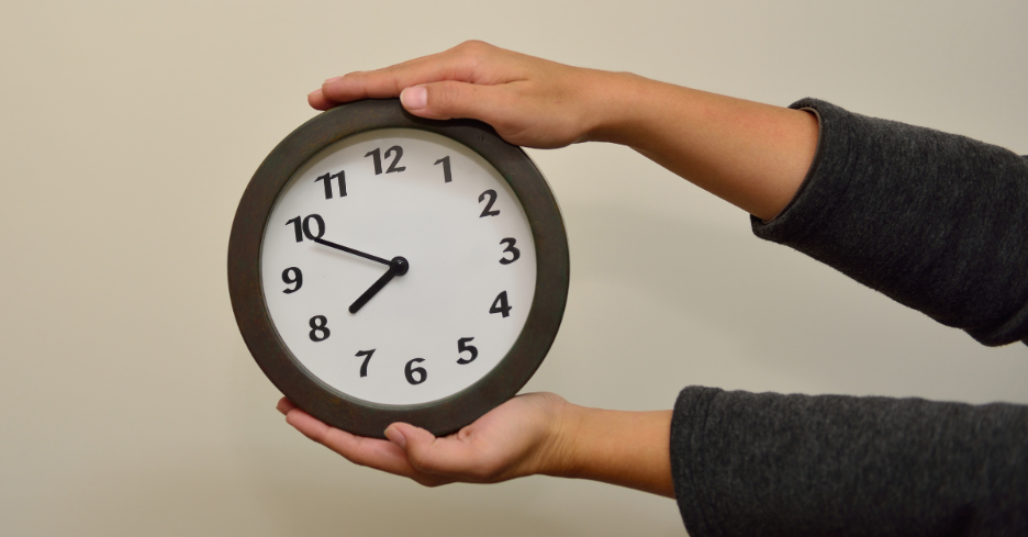 Hands holding a round clock showing the time as 7:50 against a plain background.