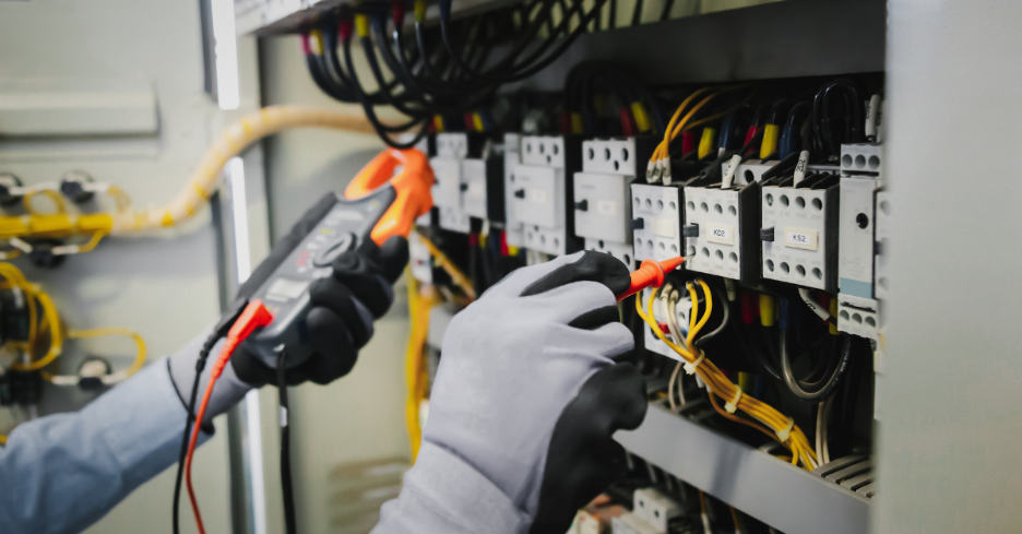 Gloved hands using a multimeter to test wiring inside an electrical control panel.