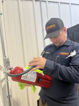 Technician in Hendrick uniform inspects a red fire extinguisher mounted on a metal wall.