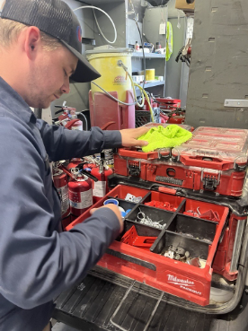 Man in cap organizing screws and tools in red Milwaukee toolboxes on truck bed in workshop.
