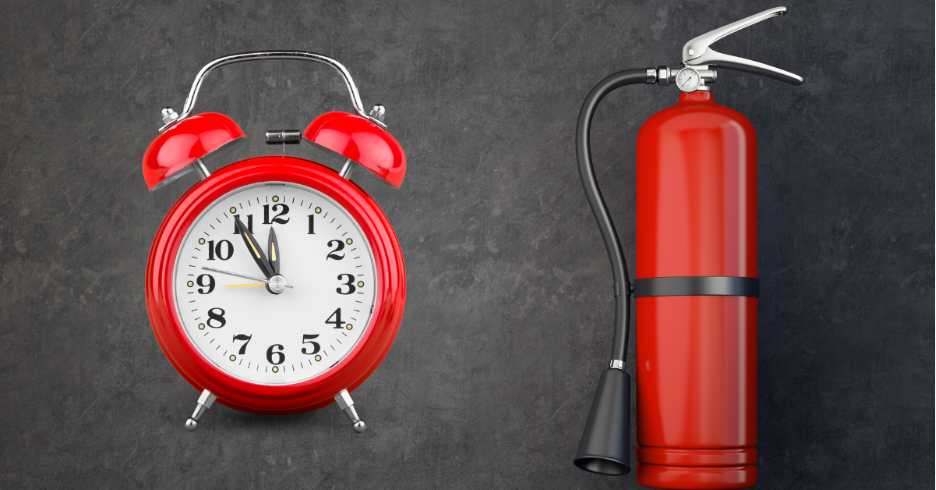 Red alarm clock showing 11:55 beside a red fire extinguisher on a gray textured surface.