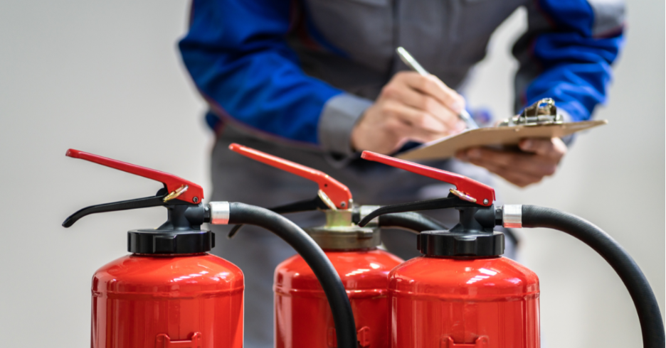 Person in uniform inspecting and writing notes on clipboard near three red fire extinguishers.