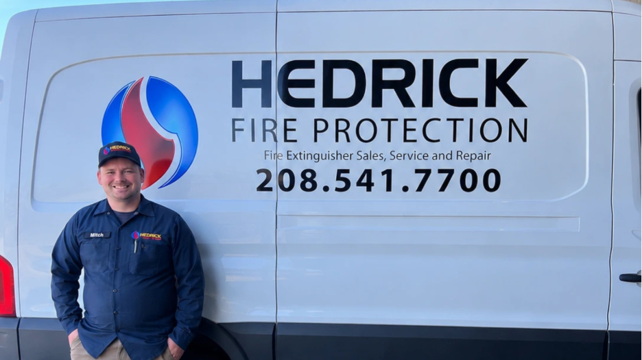 Man in navy blue work shirt and cap stands smiling by white Hedrick Fire Protection service van.