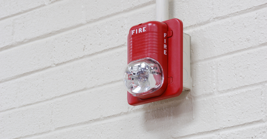 Red fire alarm with clear strobe light mounted on a white brick wall.