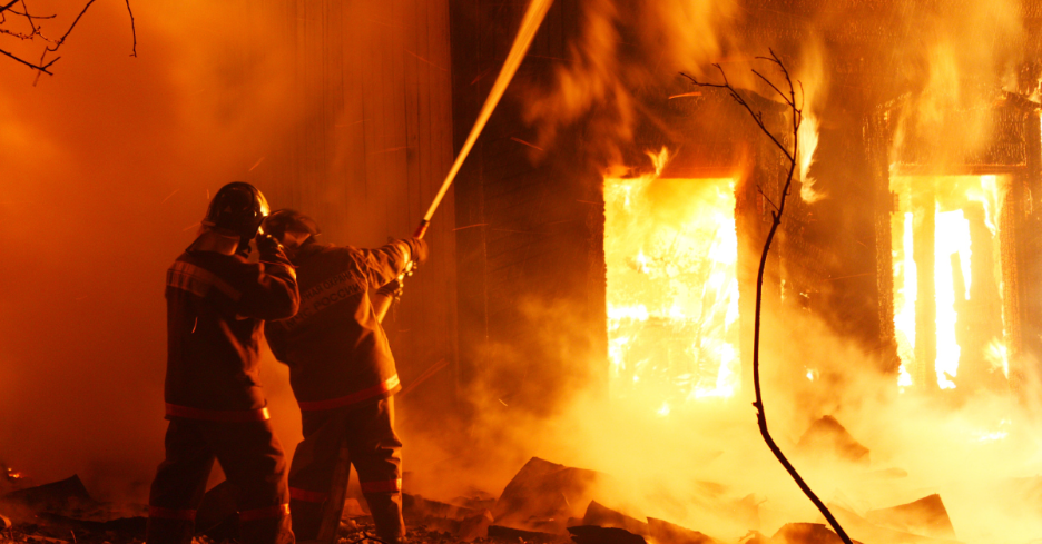 Two firefighters in protective gear spraying water on a large building engulfed in flames at night.