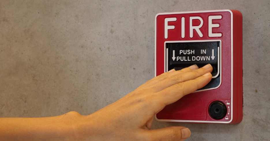 Hand reaching to press a red fire alarm pull station mounted on a concrete wall.