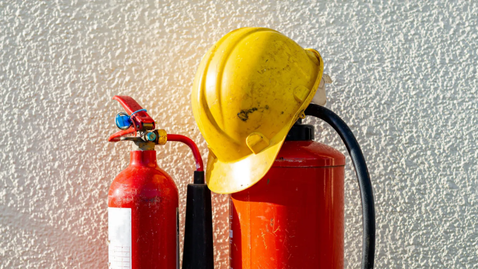 Two red fire extinguishers against a white textured wall with a yellow safety helmet resting atop one.