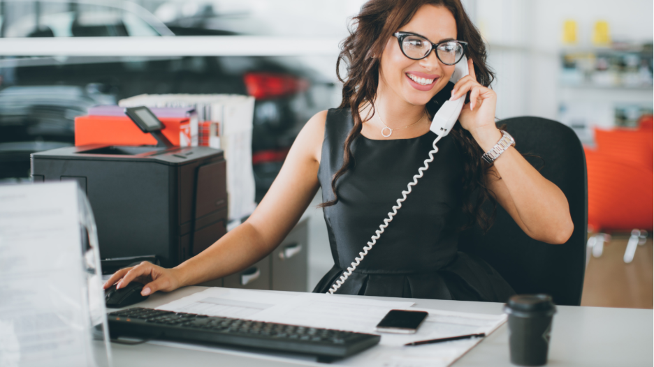 Smiling woman with glasses sitting at a desk speaking on a corded telephone while using a computer mouse with her other hand, office equipment and a blurred vehicle visible in the background.