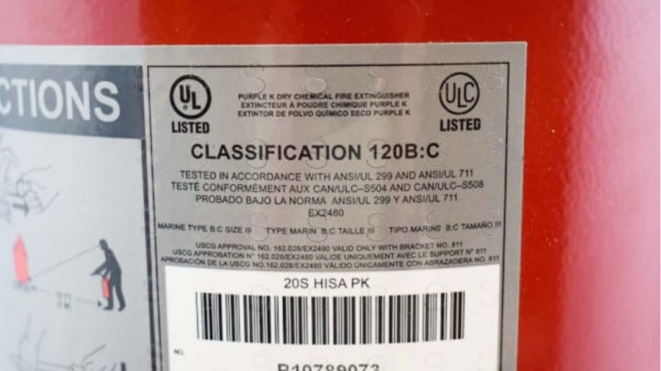Close-up of a fire extinguisher label showing classification 120B:C and UL listing marks, specifying it as a Purple K dry chemical fire extinguisher tested to ANSI and ULC standards, with approval details and a barcode below.