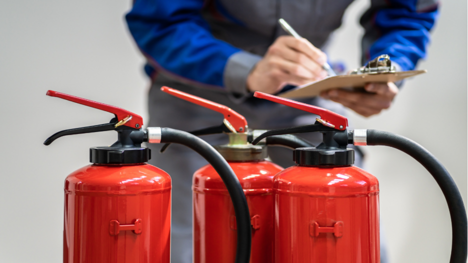 Three red fire extinguishers in the foreground with a person in blue and gray workwear writing on a clipboard in the background.