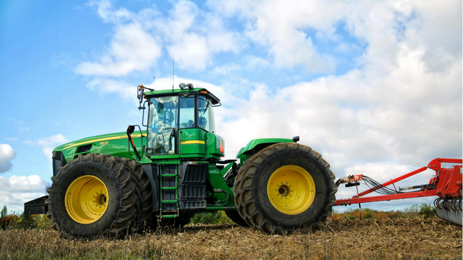 Green John Deere 9630 tractor with large yellow wheels parked on a field with cloudy sky in the background, attached to a red farming implement.